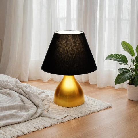 Black lamp with gold base on a wooden floor in a room with white curtains and a plant.