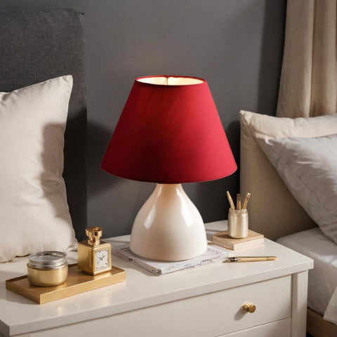 Red lampshade on a white base on a nightstand with decorative items.