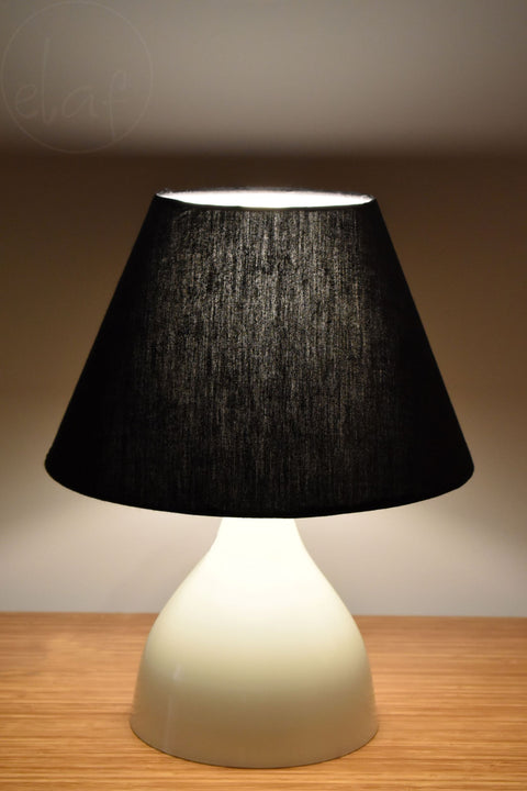 A white base table lamp with a black conical shade, illuminated, placed on a wooden surface against a dark background.