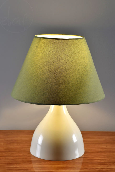 A table lamp with a white base and olive green lampshade, placed on a wooden surface.